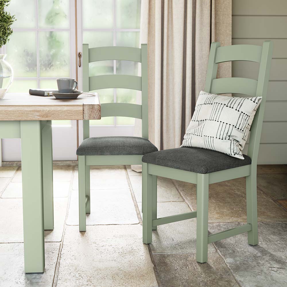 photo of two sage painted oak chairs near a matching sage painted oak dining table with charcoal grey seat cushion and a silver scatter cushion on one of the chairs. The chairs and table are set in a brightly lit kitchen with big window and cream curtains