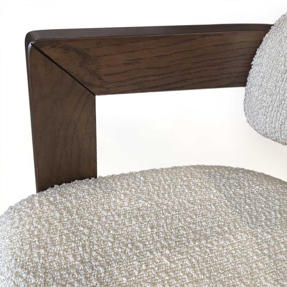 Close-up of a modern chair showing a dark brown wooden armrest with a geometric corner design and a textured white bouclé seat and back cushion.