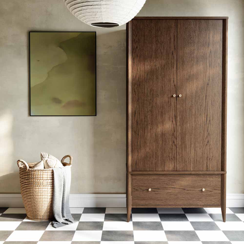 A tall two-door wooden wardrobe with a lower drawer, shown in a room with a black and white tiled floor, laundry basket and painting on the wall. The cabinet is made of light oak wood with a natural grain and features small round metal knobs.