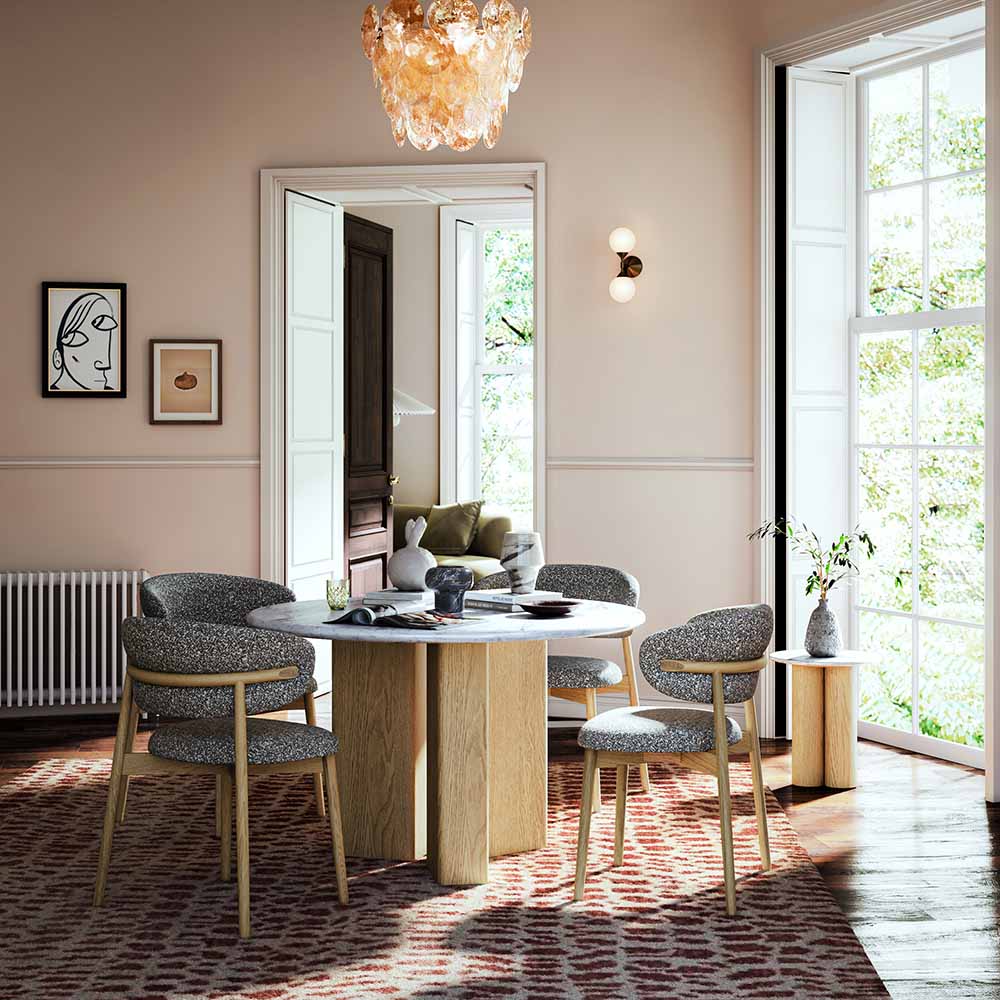 A round marble top dining table with a light wood pedestal base and four upholstered dining chairs with gray textured fabric and light wood frames, arranged in a bright, modern dining room with a large window, patterned rug, and wall sconces.