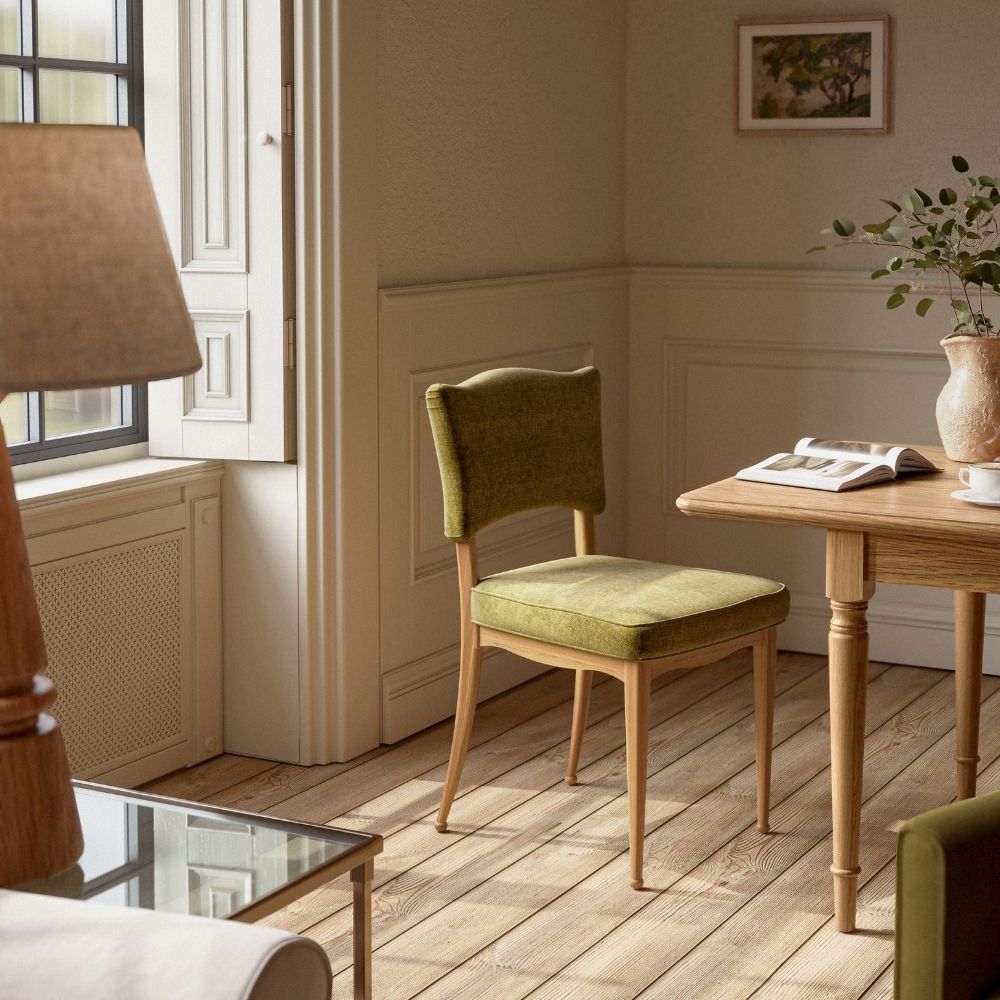 An upholstered dining chair with an olive green fabric seat and backrest, set in a warm, neutral-toned room next to a wooden table.
