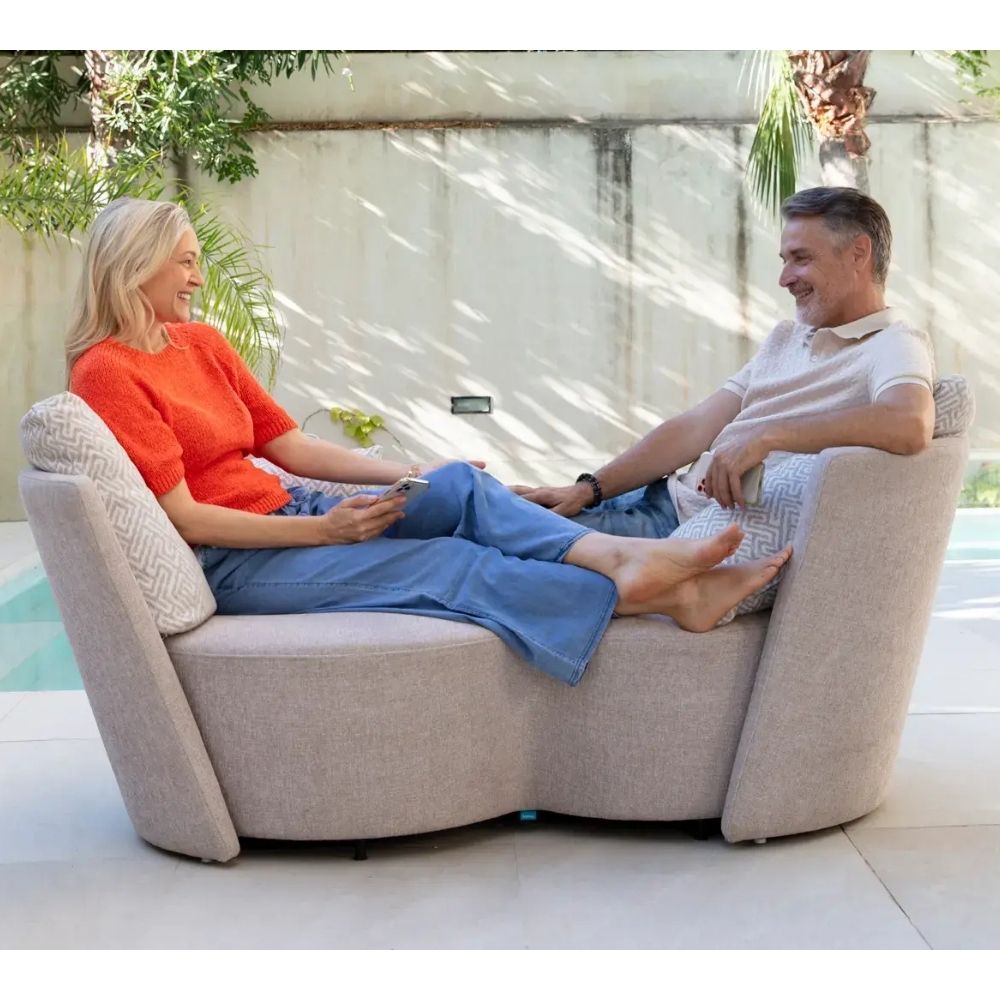 Two people sitting on a large beige infinity sofa with a pool and garden in the background.