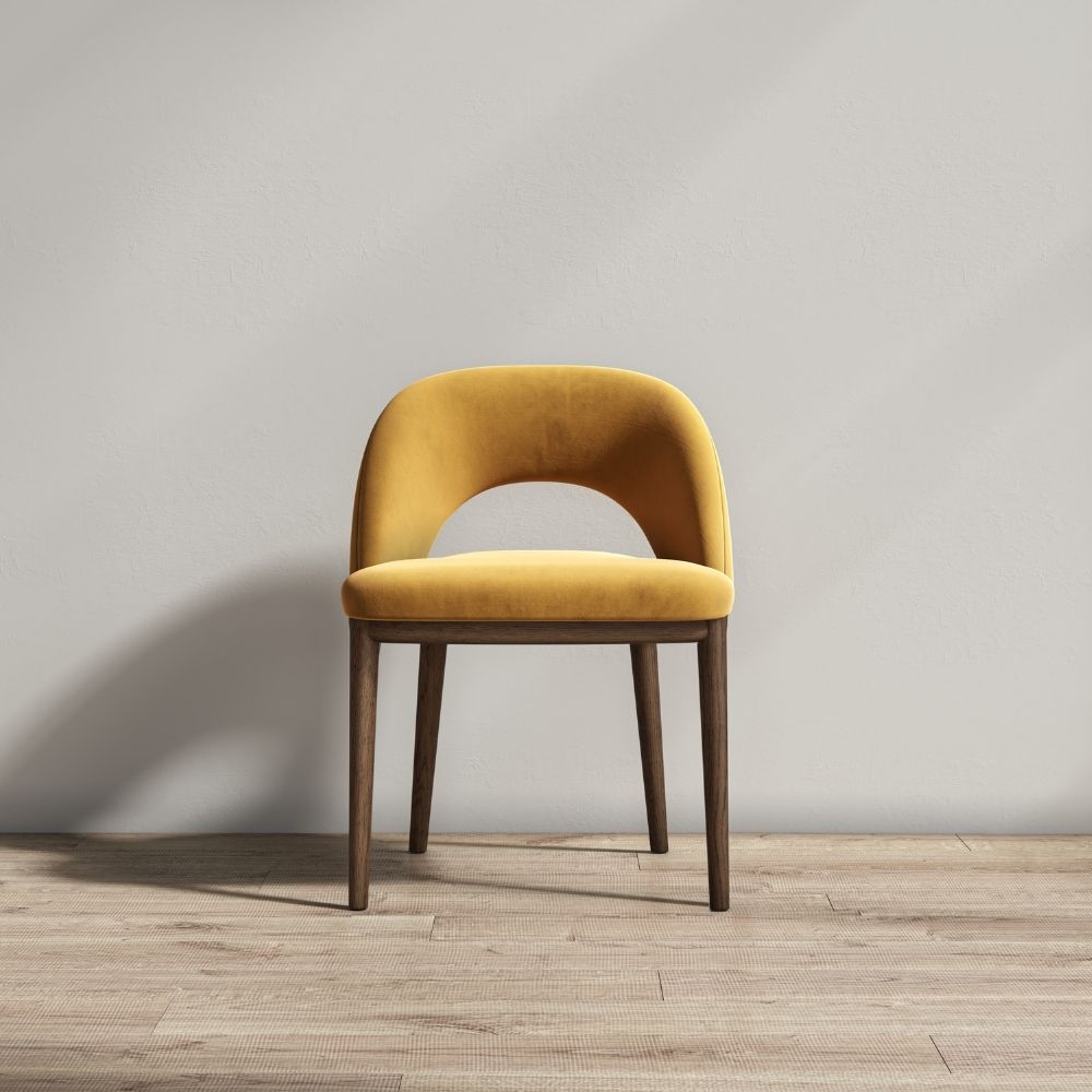 A modern accent chair with a curved mustard yellow velvet seat and backrest, supported by four tapered dark brown wooden legs, set against a neutral gray wall and light wood floor.