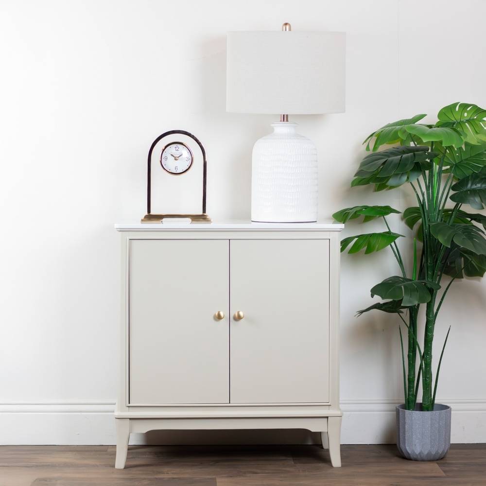 Stone / taupe / beige cabinet with gold handles, lamp, and clock against a white wall with a plant on the right.