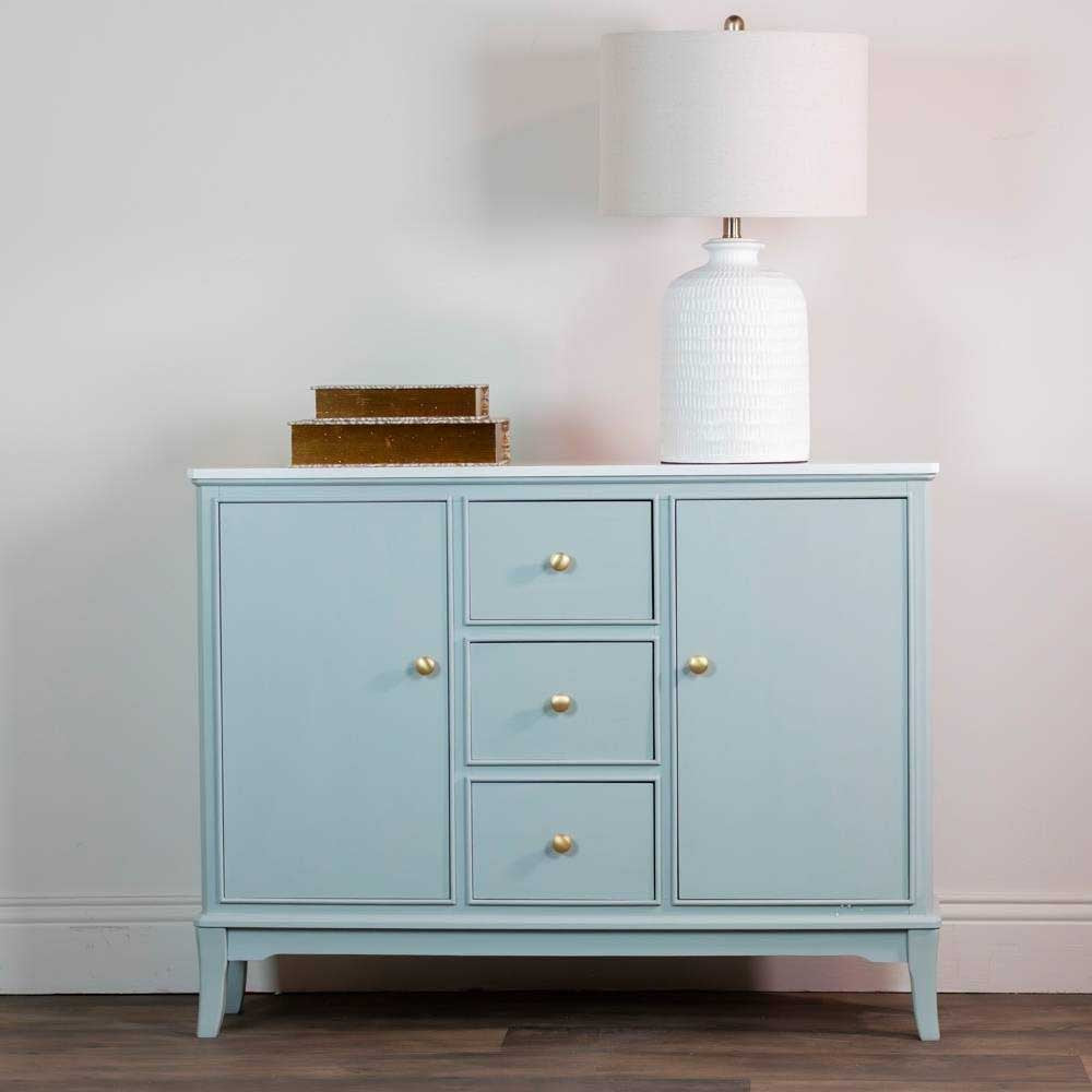 A sage green wooden sideboard with three drawers in the center and two doors on the sides, accented with small round gold knobs, decorated with a lamp and two books, shown against a plain white background.