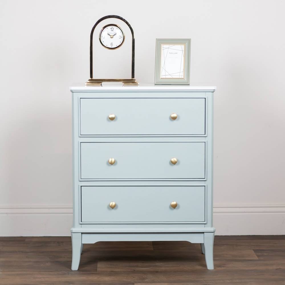 A three-drawer chest of drawers in a light sage green finish with round gold knobs, styled on a dark wood floor against a white wall with decorative objects on top.