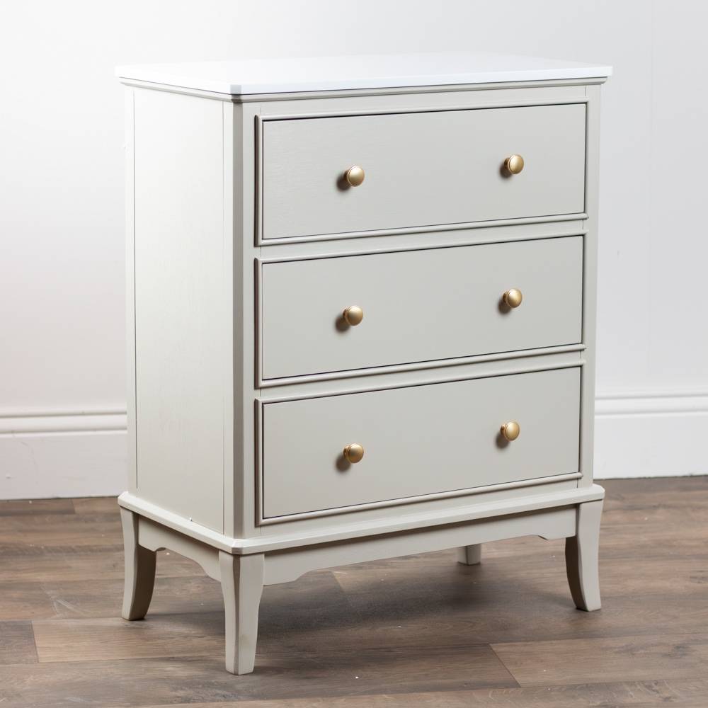 A three-drawer chest in a light beige / stone coloured finish with small round gold knobs, shown standing on a wood floor against a white wall.