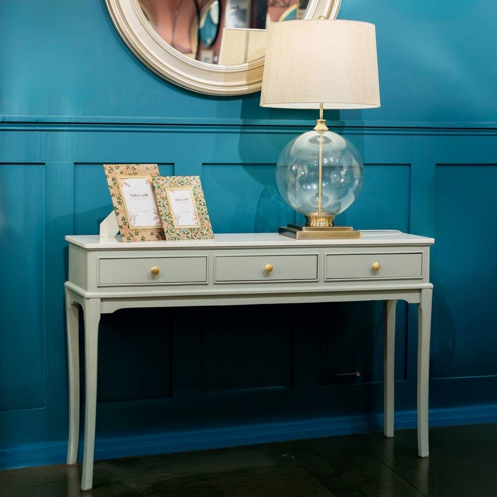 Sage green console table with a lamp and framed pictures against a blue wall.