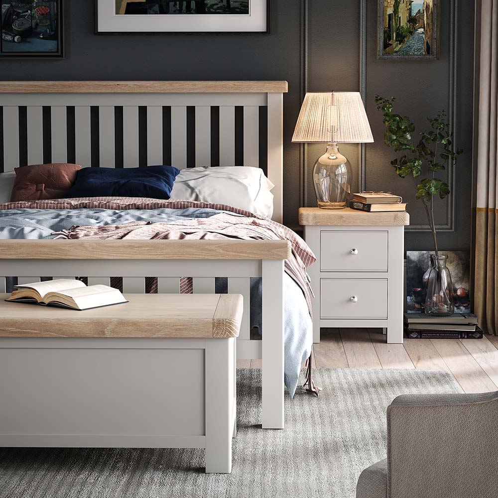 Lifestyle photo of a grey double bed in bedroom with matching sage bedside locker, and blanket box/ There are photos on the wall and a lamp on the bedside table.