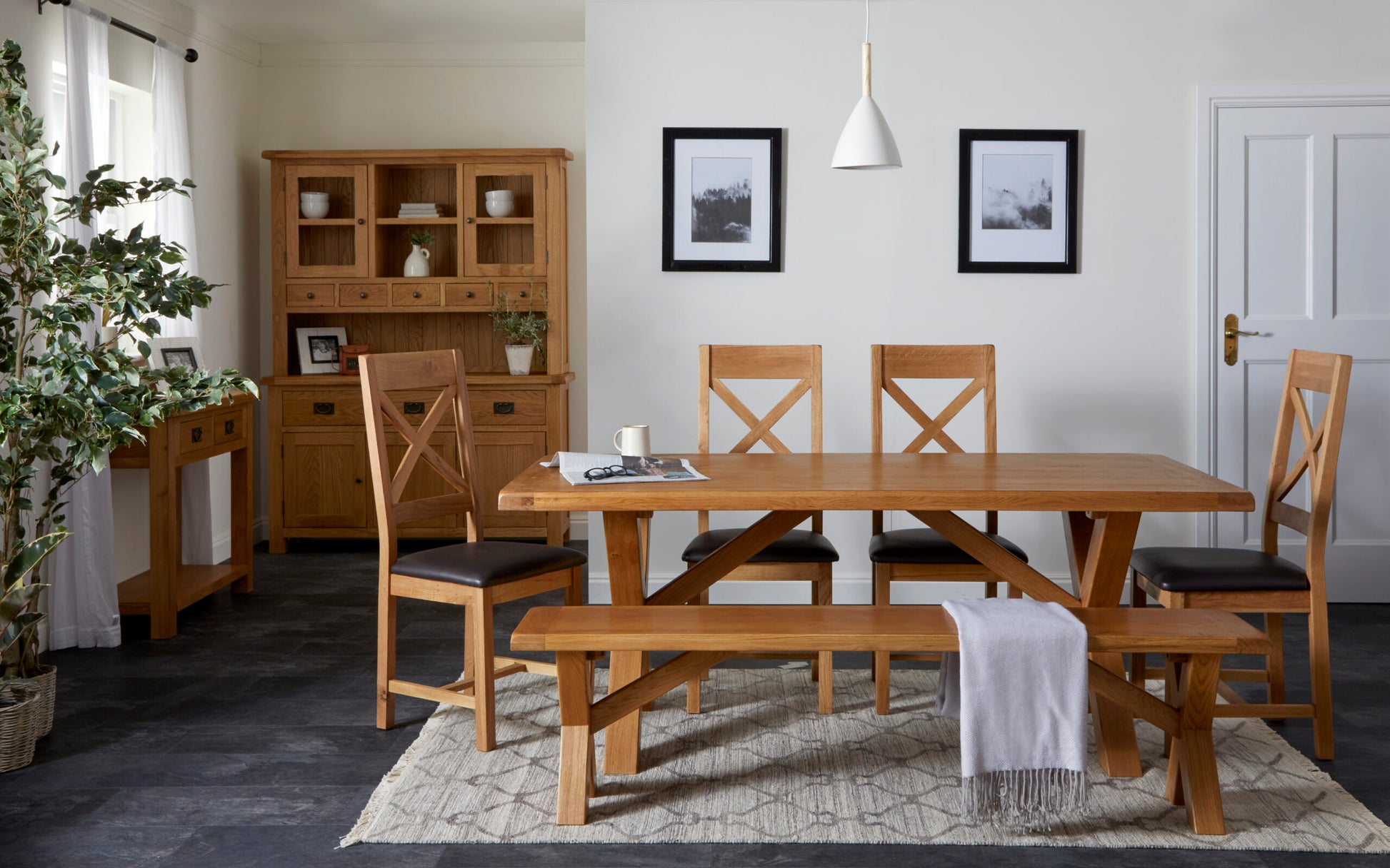 lifestyle image of the oak cross back chairs placed around a table in a homely kitchen. 2 chairs are on the far side of the table facing the viewer. One chair is at an angle on the left hand side as if it had been pulled out by the person sitting at the head of the table.