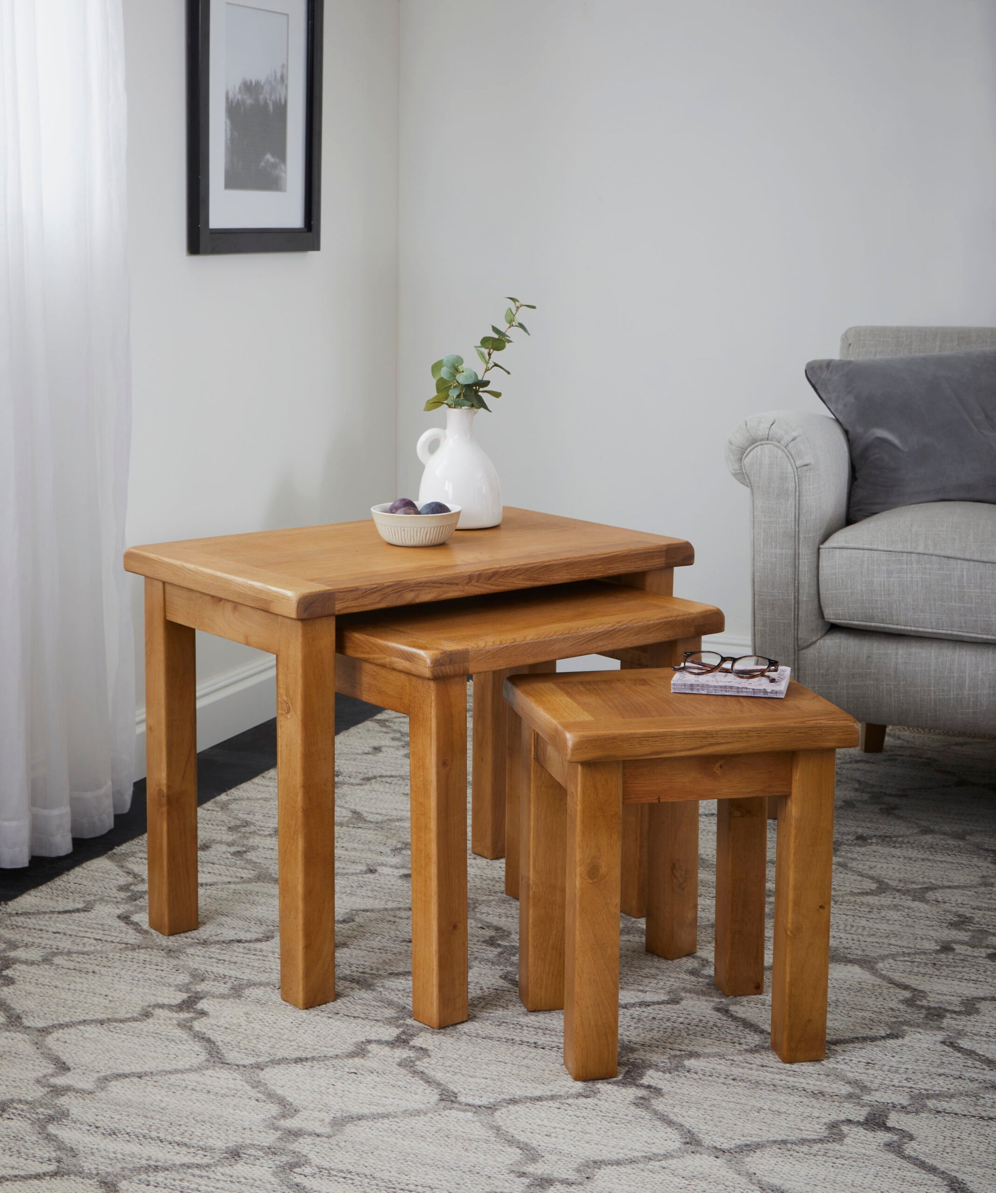 lifestyle image of a nest of three oak tables with a vase and decorative bowl on top, set in a sitting room on a rug next to a grey couch.