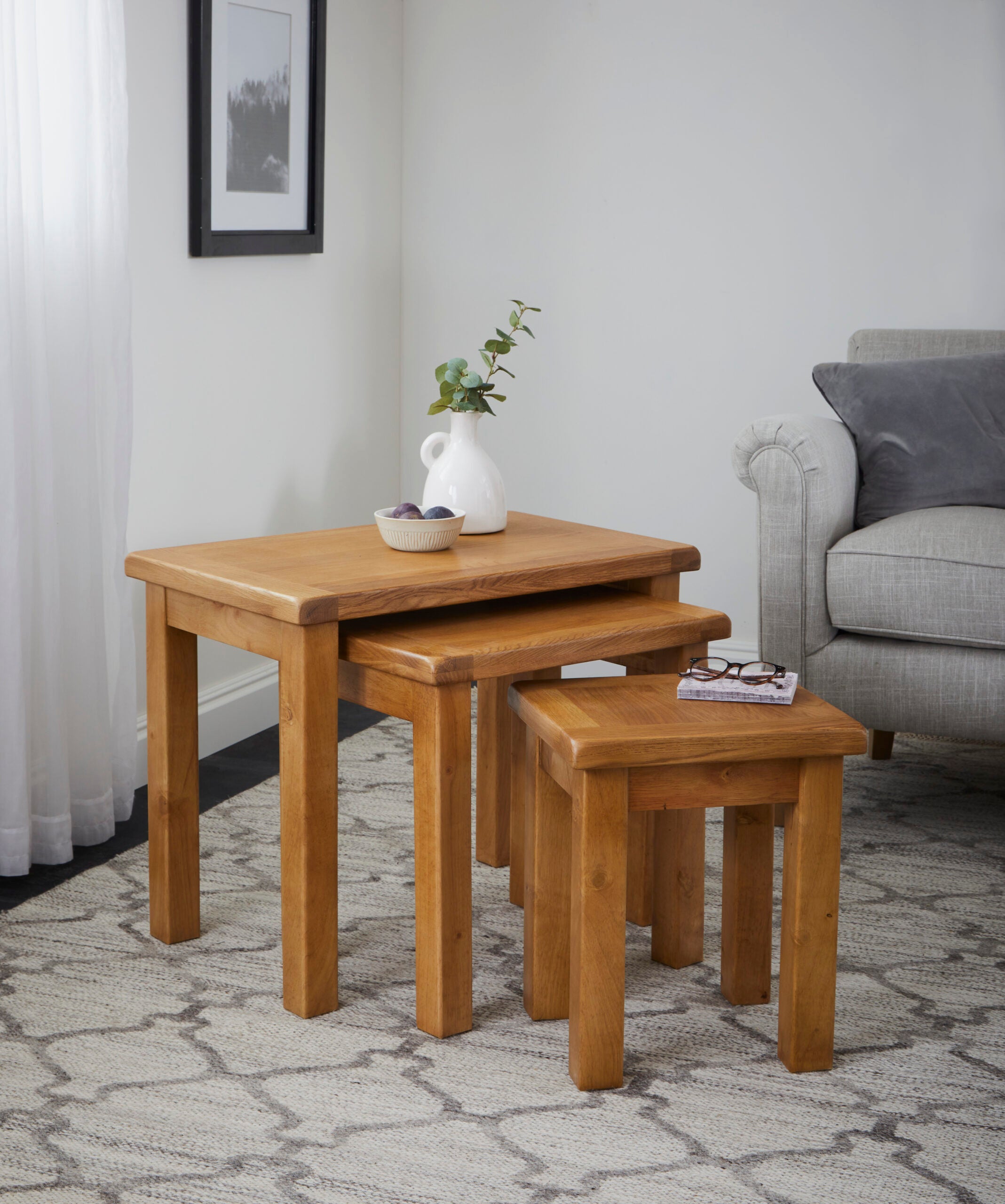 lifestyle image of a nest of three oak tables with a vase and decorative bowl on top, set in a sitting room on a rug next to a grey couch.
