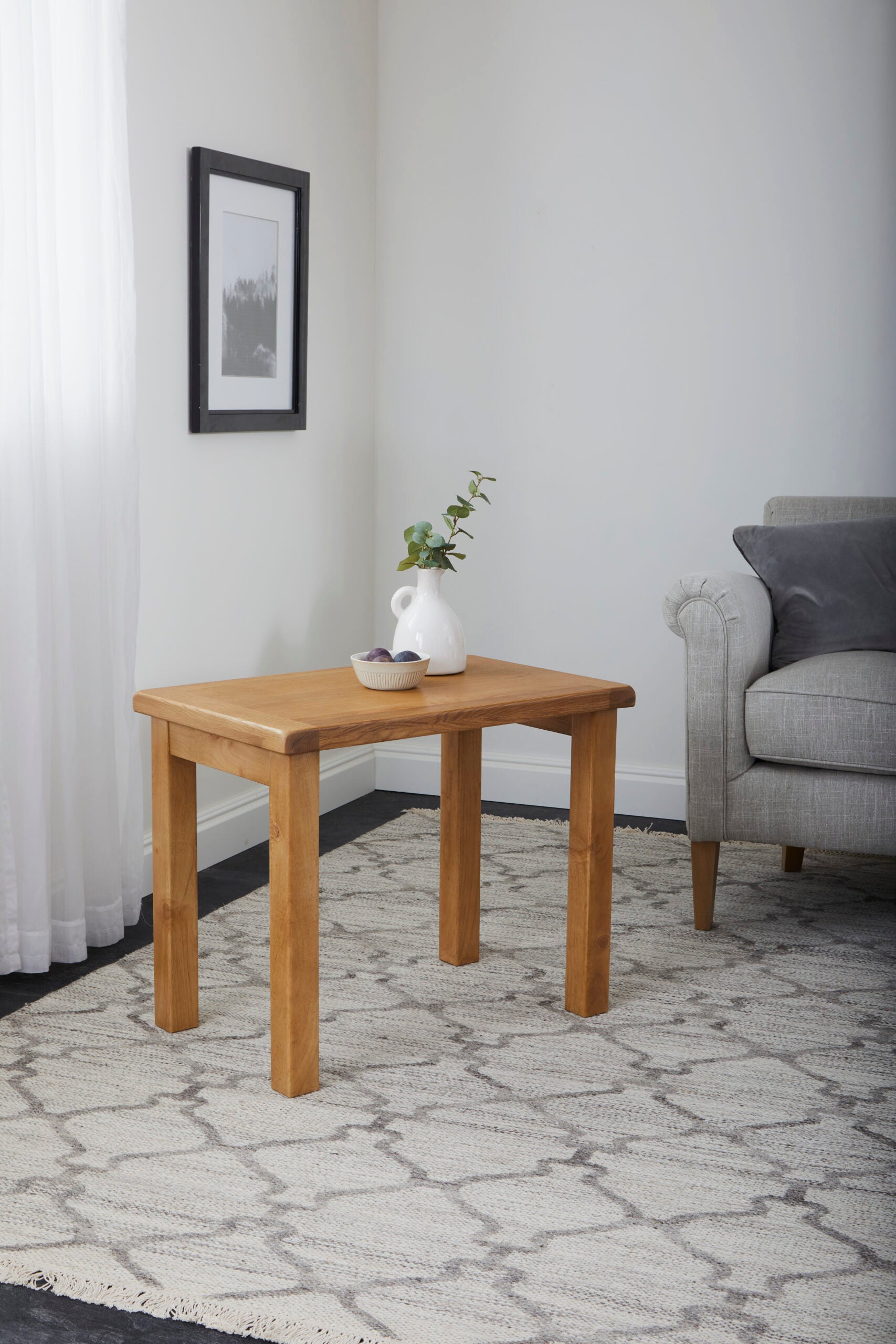 1 table of a nest of three oak tables with a vase and decorative bowl on top on a rug next to a couch.