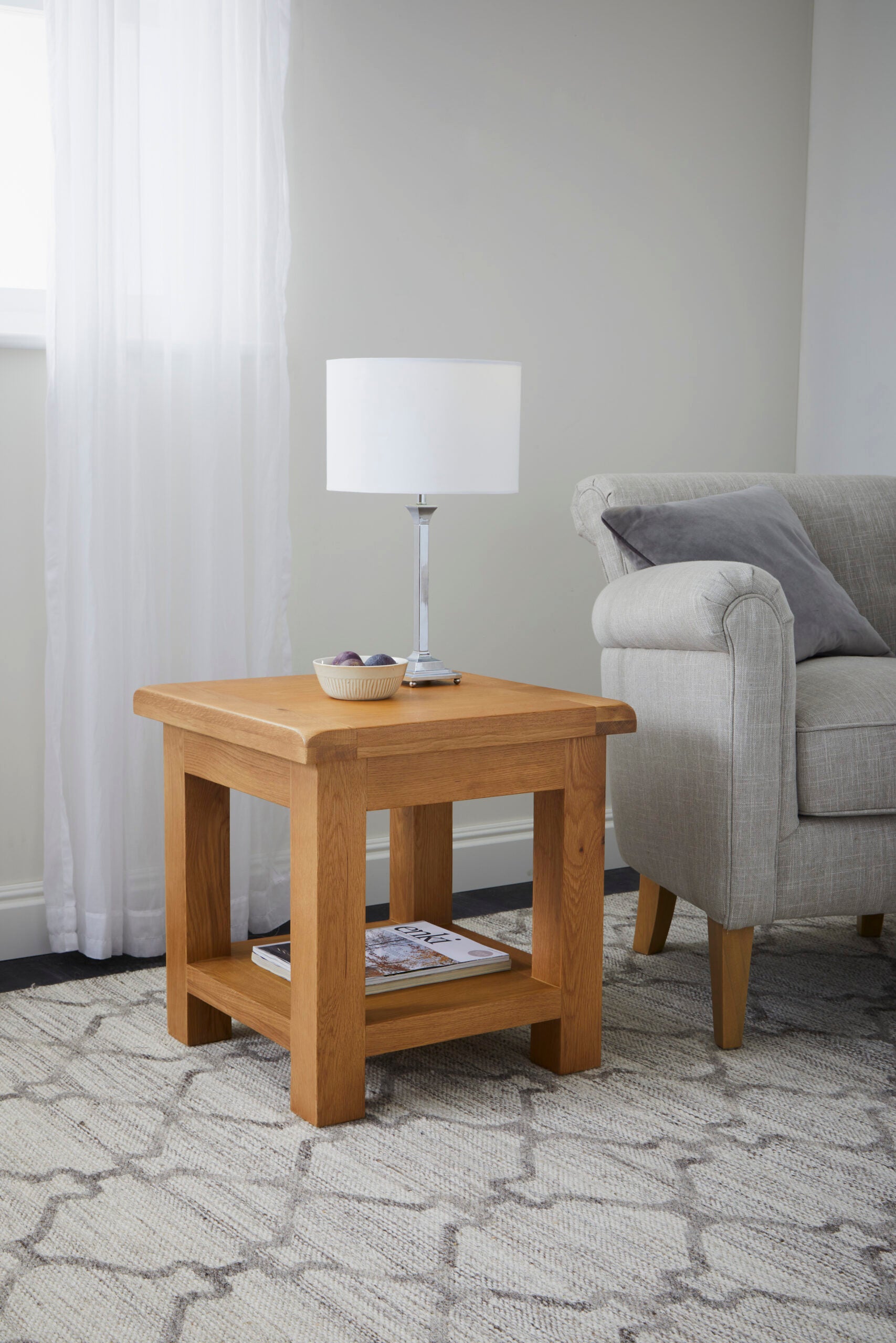 lifestyle image of oak square lamp table in sitting room next to a grey couch with a white lamp and smal decorative bowl on the tabletop and a magazine on the low open storage shelf underneath.