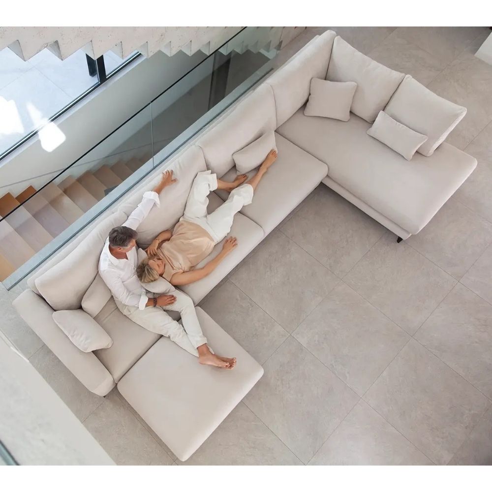 Two people lying on a large beige Fama sectional sofa in a modern living room.