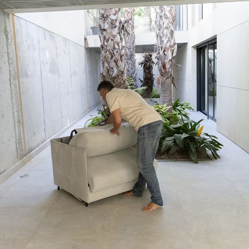 Person unfolding single sofa bed in a modern indoor setting with plants and concrete walls.