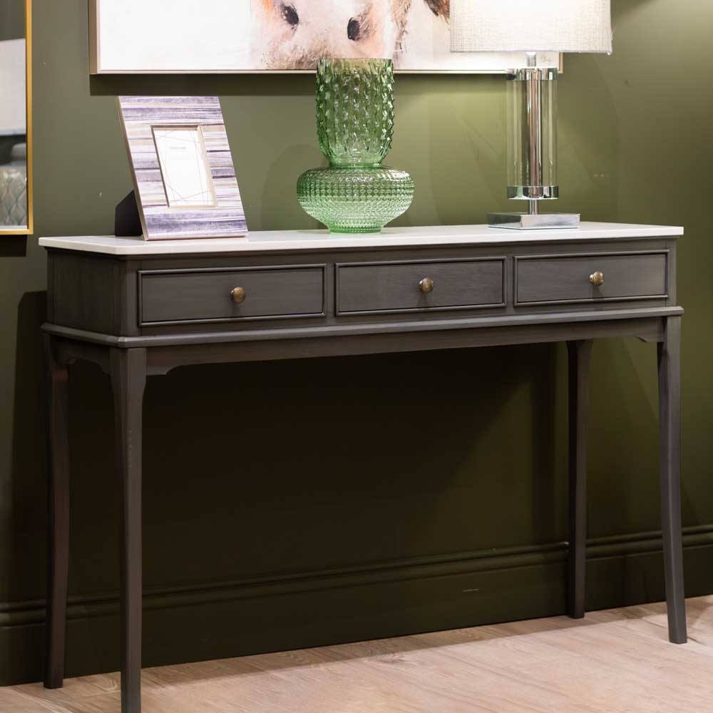 A dark brown coloured console table with three drawers and gold knobs, styled with a picture frame, a green galss vase, and a lit table lamp against a moss green wall.