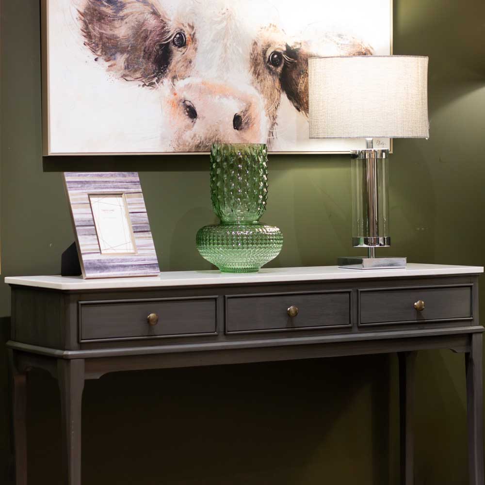 A dark brown console table with three drawers and brass knobs, featuring a white marble top. The table is styled with a green textured glass vase, a striped picture frame, and a modern table lamp, set against an olive green wall with a cow painting hanging above.