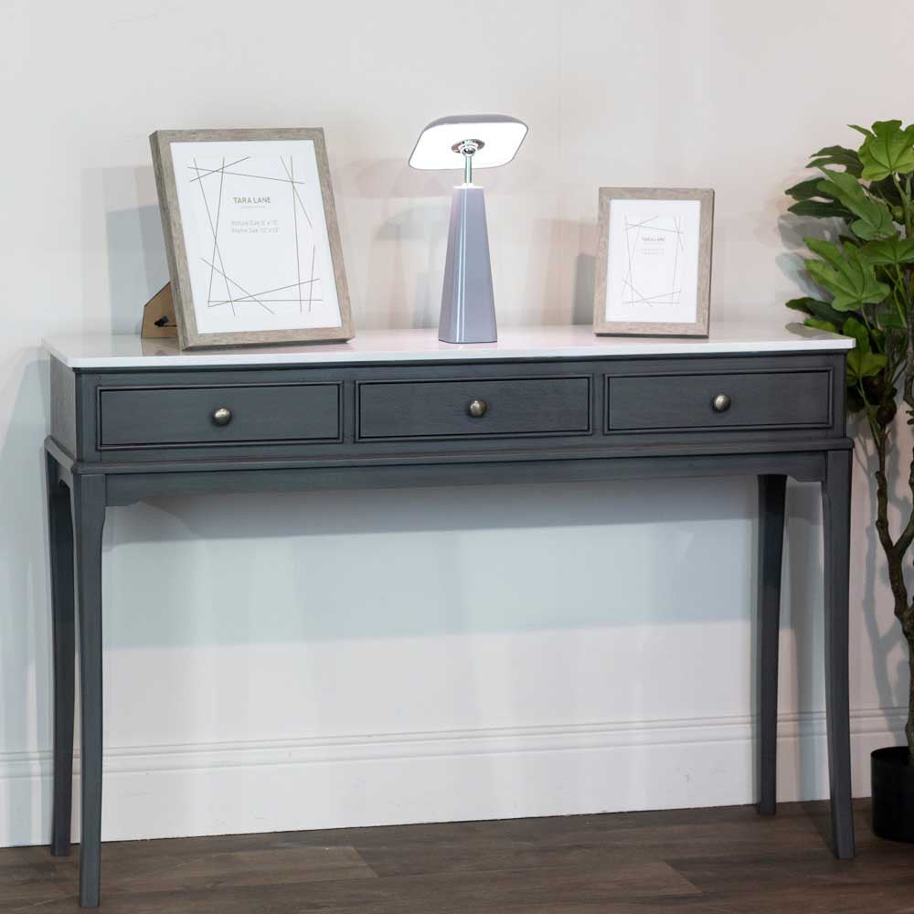 A dark brown coloured console table with three drawers and gold knobs, styled with two picture frames, and a  small LED mushroom lamp, against a neutral wall.