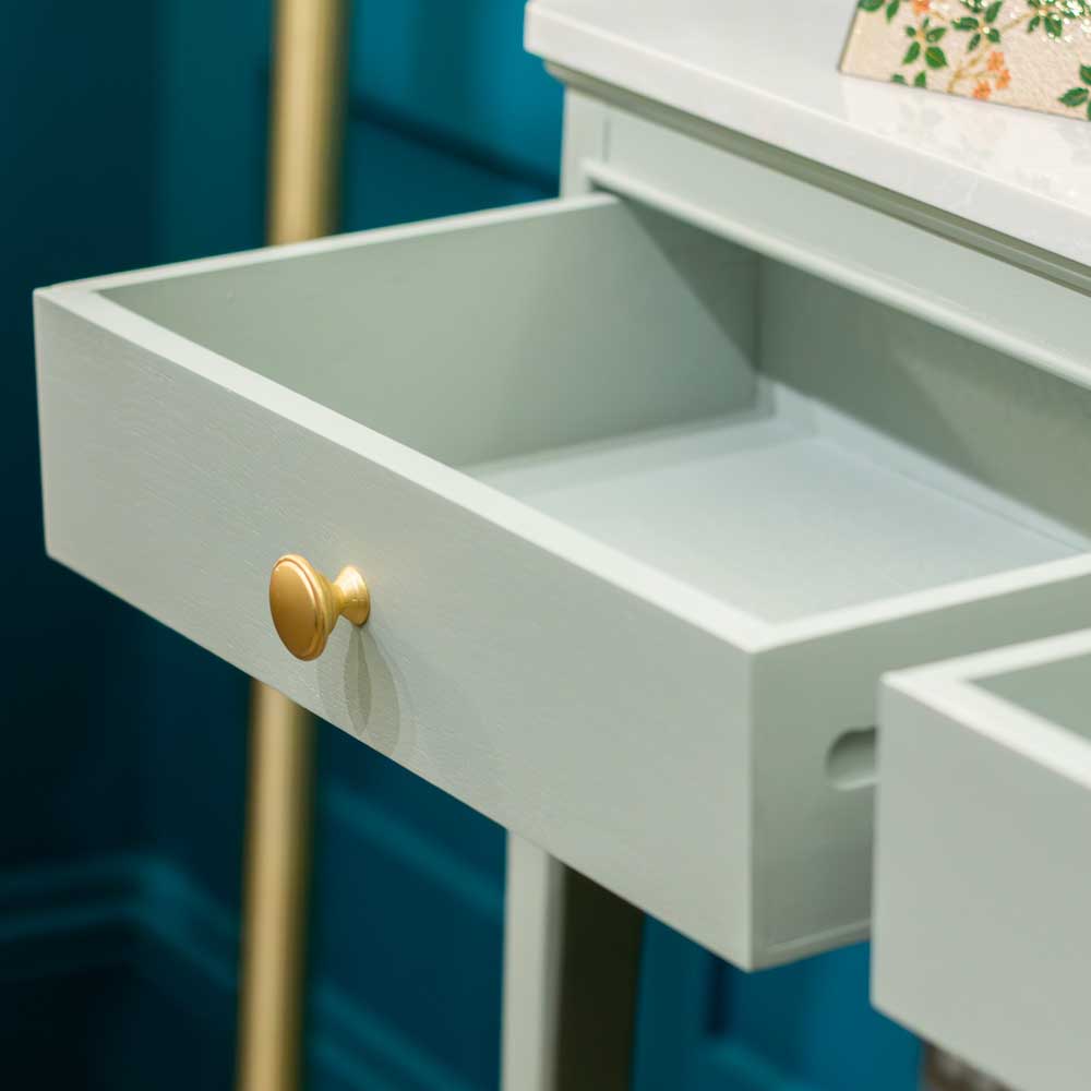 A close-up of a light sage green drawer with a single round gold pull knob, shown partially open against a dark teal background.