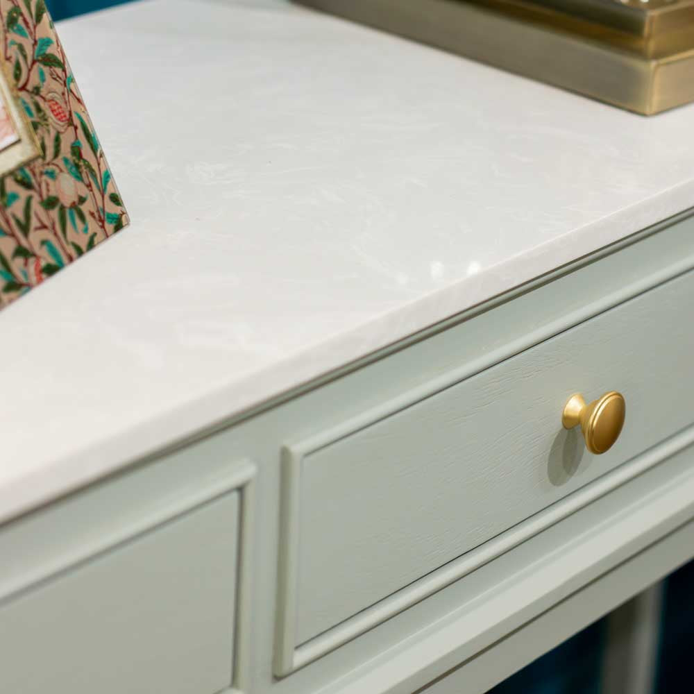 Close-up of a white marble-look countertop with a light sage green console base and a round gold drawer knob.