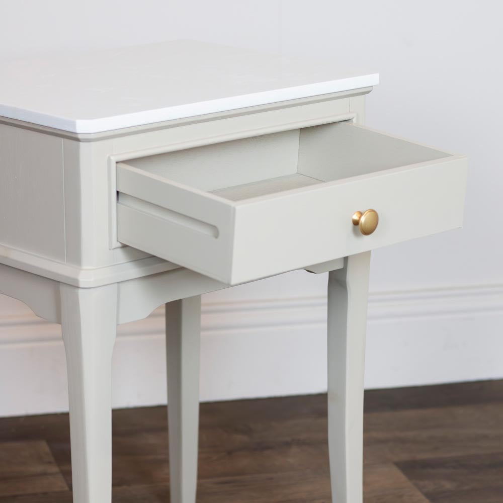 Taupe / beige lamp / accent table with an open drawer featuring a round gold pull knob, set against a white wall and dark wood floor.
