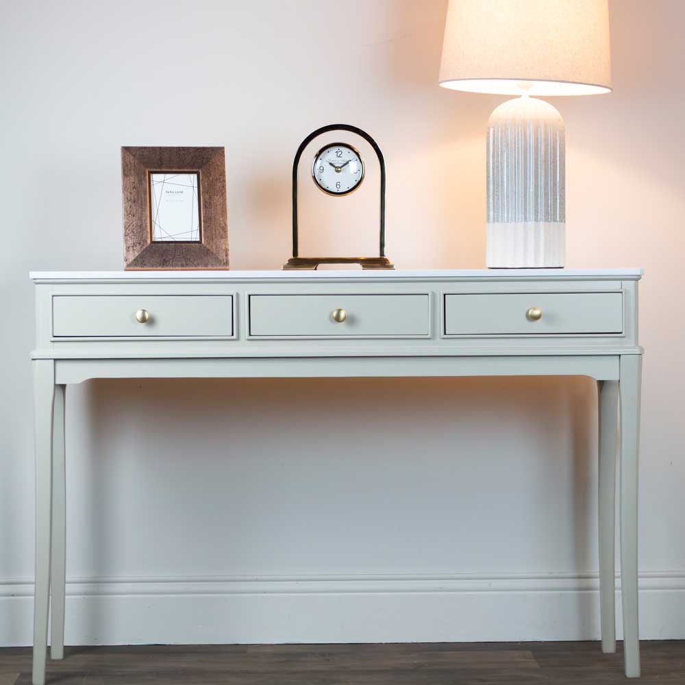 A beige / stone coloured console table with three drawers and gold knobs, styled with a picture frame, a small arch clock, and a lit table lamp against a neutral wall.