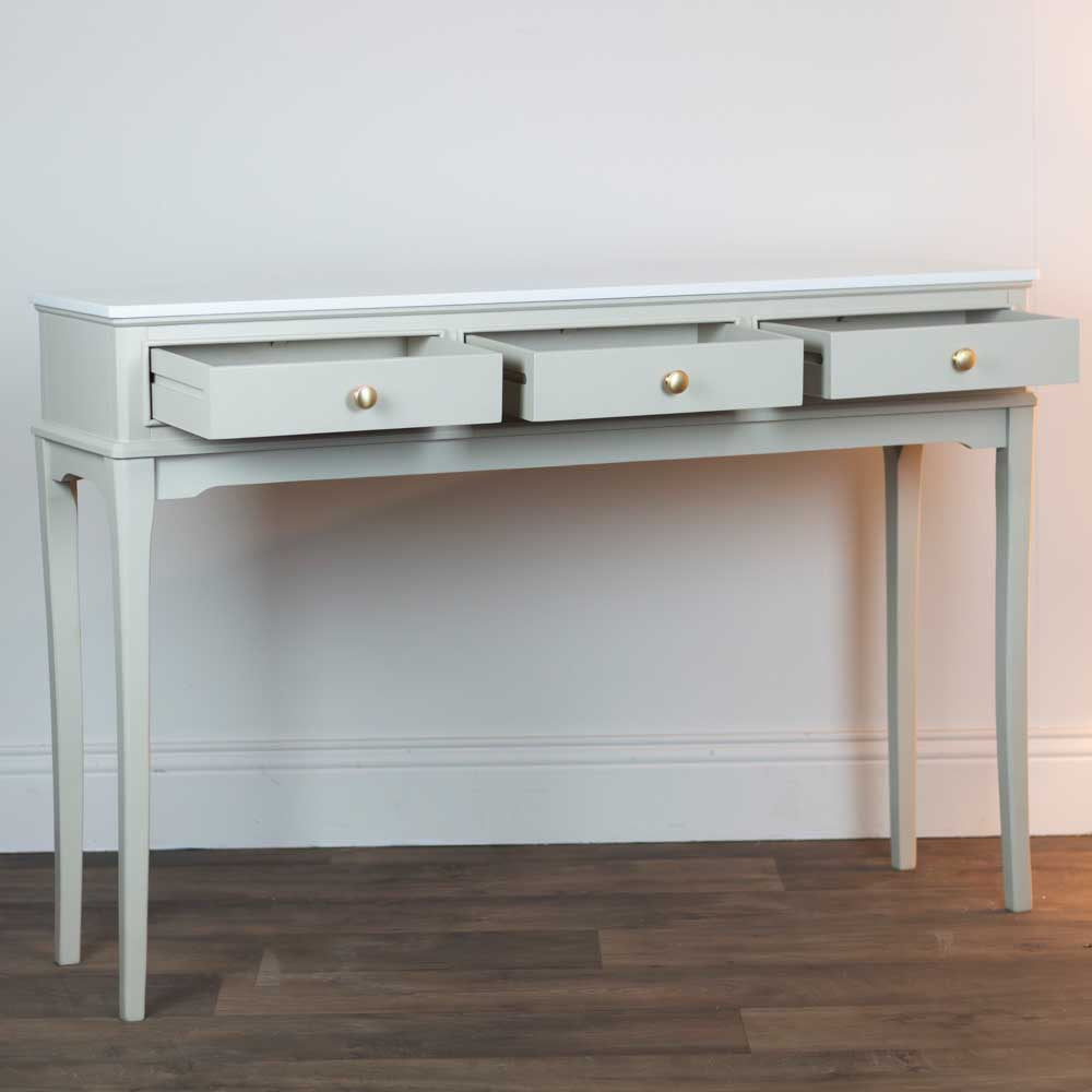 Light beige / stone coloured console table with three drawers on a wooden floor against a white wall.