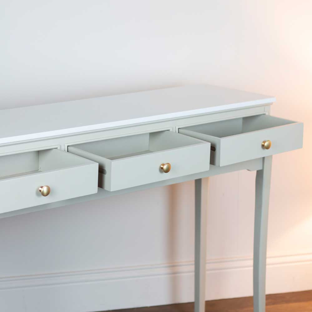 A beige / stone coloured console table with three open drawers and small round gold knobs, shown against a white wall with a warm light on the right side.