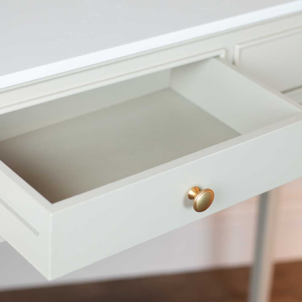 A close-up of a beige / stone coloured drawer pulled open on a white desk, featuring a round gold metal knob.