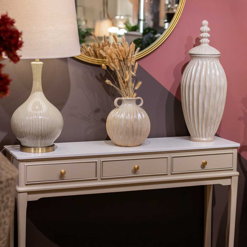 A beige / stone coloured console table with three drawers and gold knobs, styled with a vase, table lamp and decorative vessel, with a  mirror in the background, up aagainst a pink and purple painted wall.