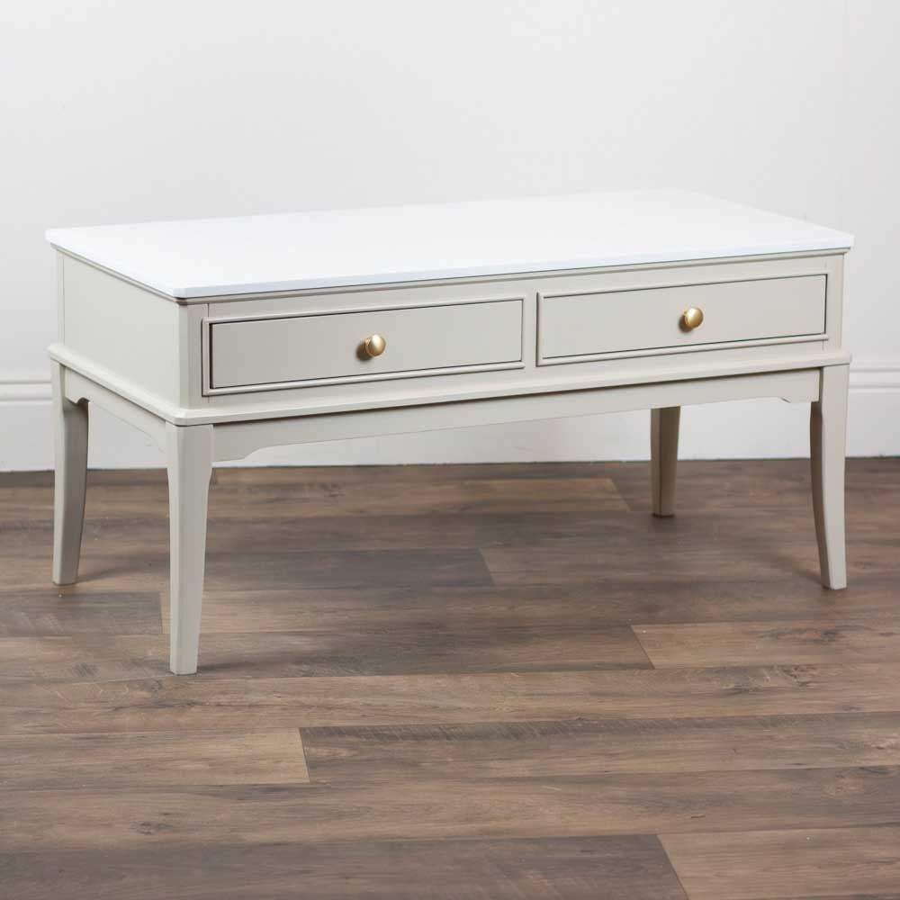 A light beige / stone coloured coffee table with two drawers and small round gold knobs, shown against a plain white background.