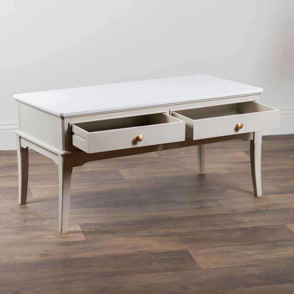 A light beige / stone coloured coffee table with two open drawers, featuring small round gold knobs, set against a neutral wall and dark wood flooring.
