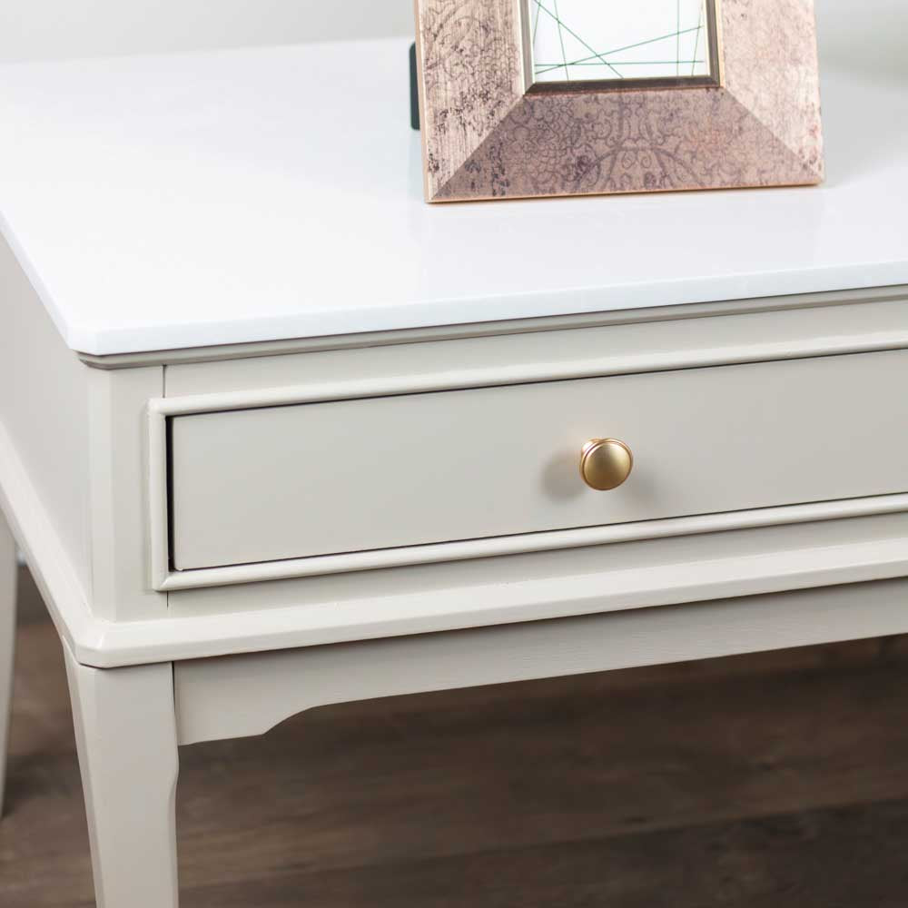 A close-up of a light beige / stone coloured coffee table with a single drawer visible, featuring a round gold pull knob and a glossy white top surface.