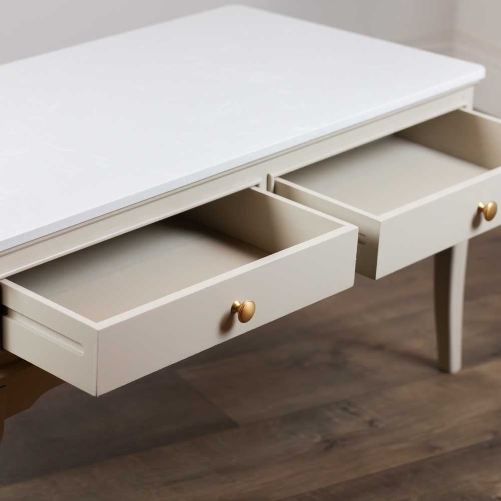 A light beige / stone coloured coffee table with two open drawers and small round gold knobs, set against a dark wood floor.