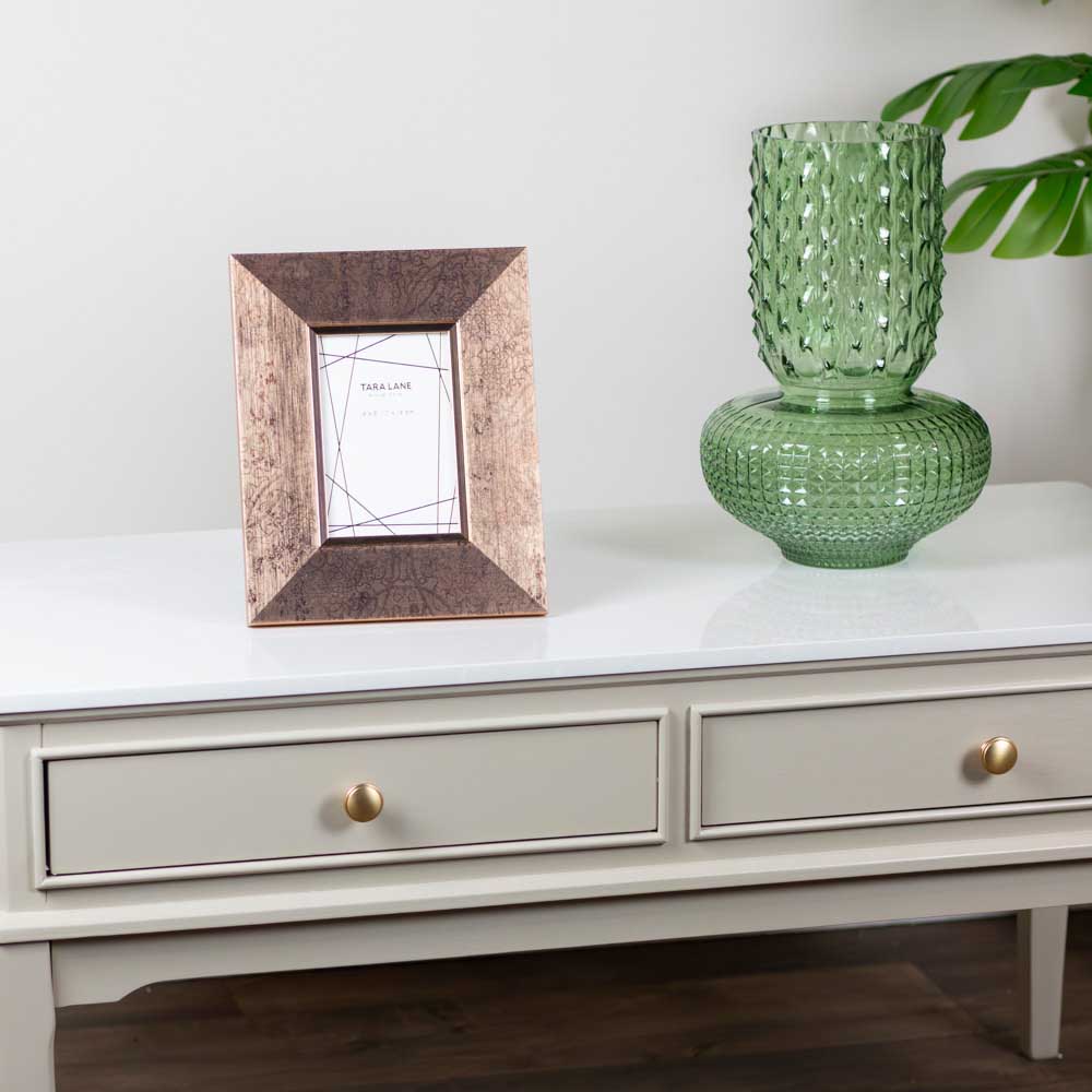 A square picture frame with a distressed brown wood-grain finish sits on a light beige / stone coloured coffee table next to a green textured glass vase. The background is a clean white wall with a green plant leaf visible in the upper right corner.