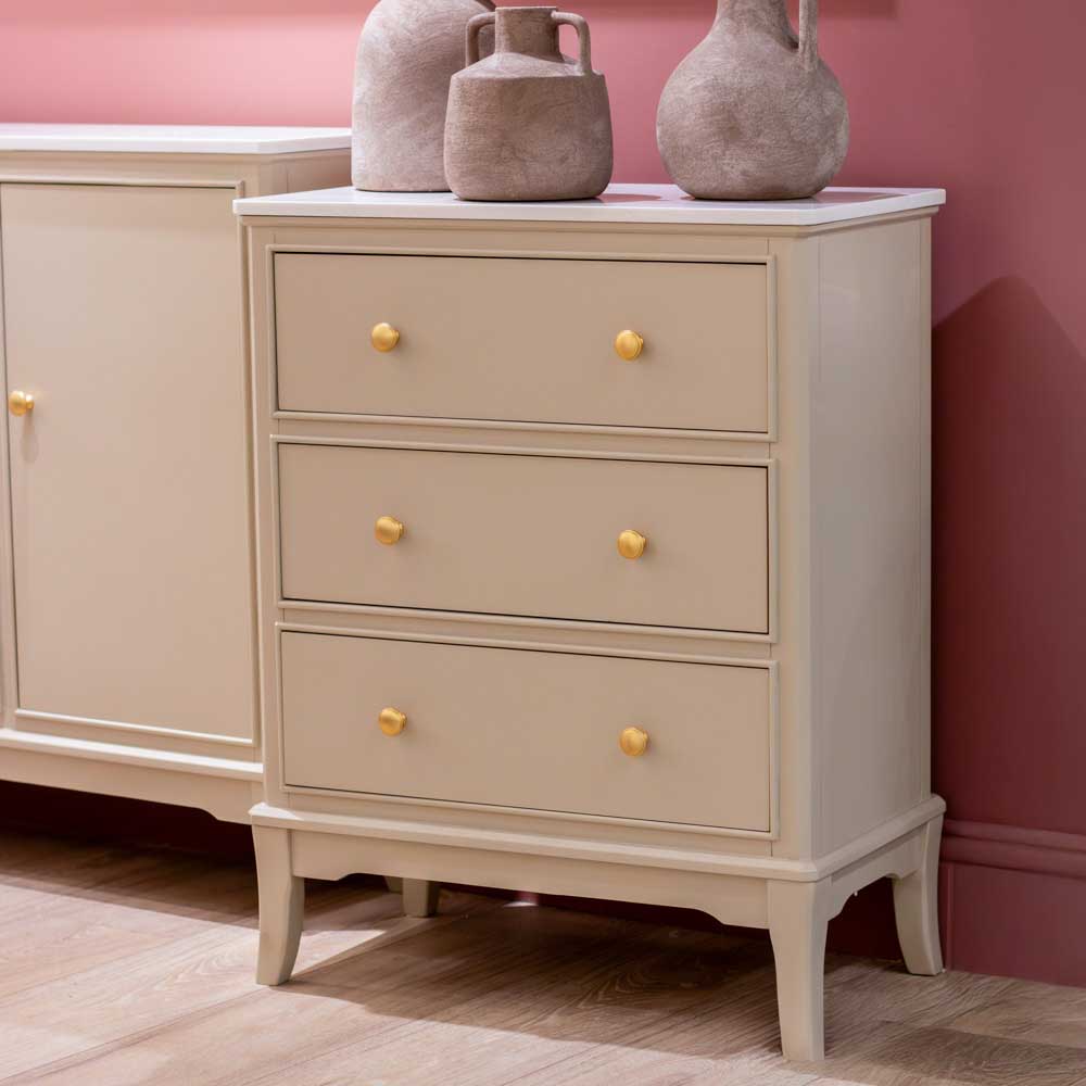 A three-drawer chest in a light beige / stone coloured finish with a white marble top, decorated with three rustic ceramic vases, set against a dusty rose wall and light wood flooring.