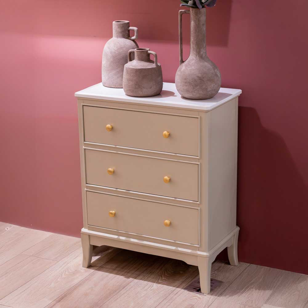 A three-drawer chest of drawers in a light beige / stone coloured finish with a white marble top, decorated with three rustic ceramic vases, set against a dusty rose wall and light wood flooring.