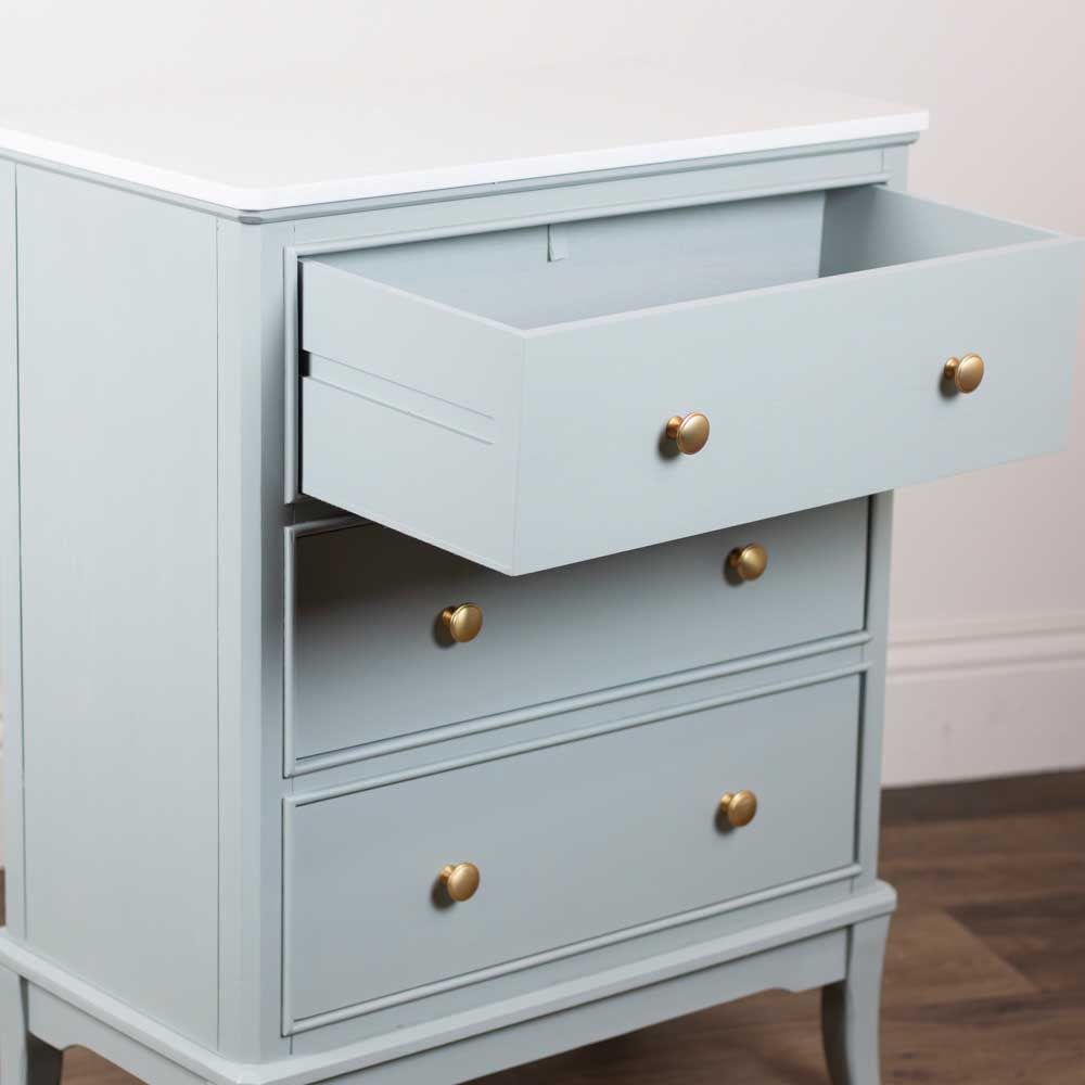 A sage green three-drawer dresser with brass knobs, shown with the top drawer pulled open, set against a neutral indoor background.