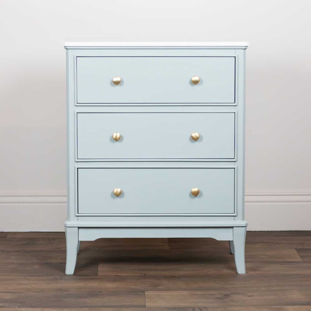 A three-drawer chest of drawers in a light sage green finish with small round gold knobs, standing on a dark wood floor against a white wall.