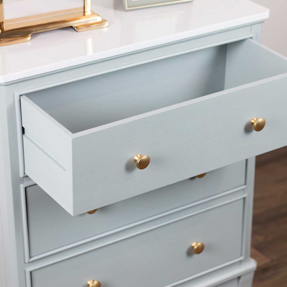 A close-up of a sage green wooden dresser with one drawer pulled open, featuring round gold knobs, set against a white tabletop and a blurred wood floor background.