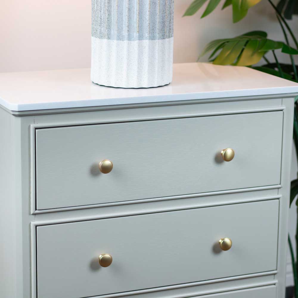 A light sage green three drawer dresser with round gold knobs, shown with a white ceramic vase on top against a soft neutral background with a green plant leaf visible on the right. 