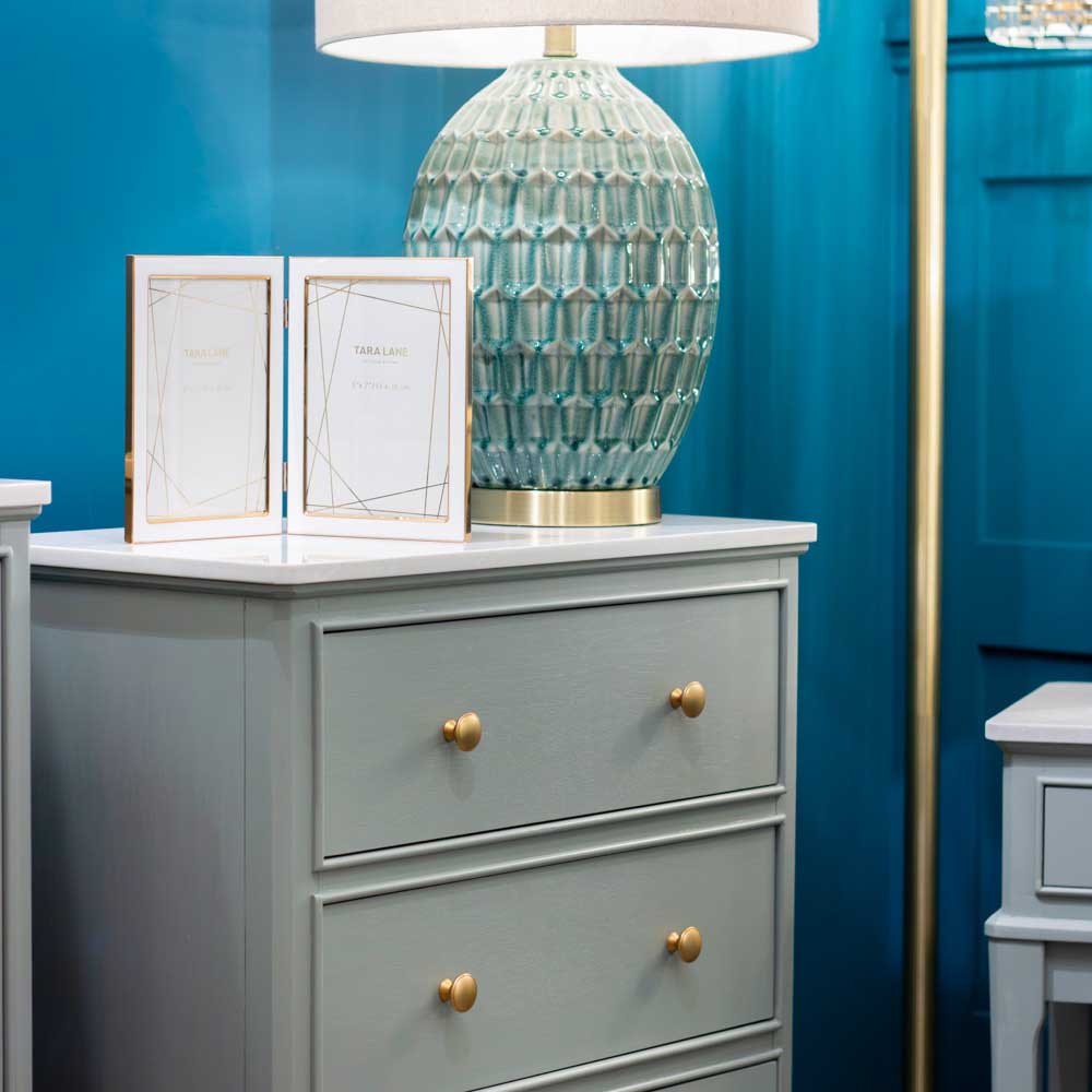 A sage green three-drawer dresser with gold knobs in a styled room setting, topped with a textured light blue-green table lamp, two picture frames, and a white lampshade, against a deep blue paneled wall.