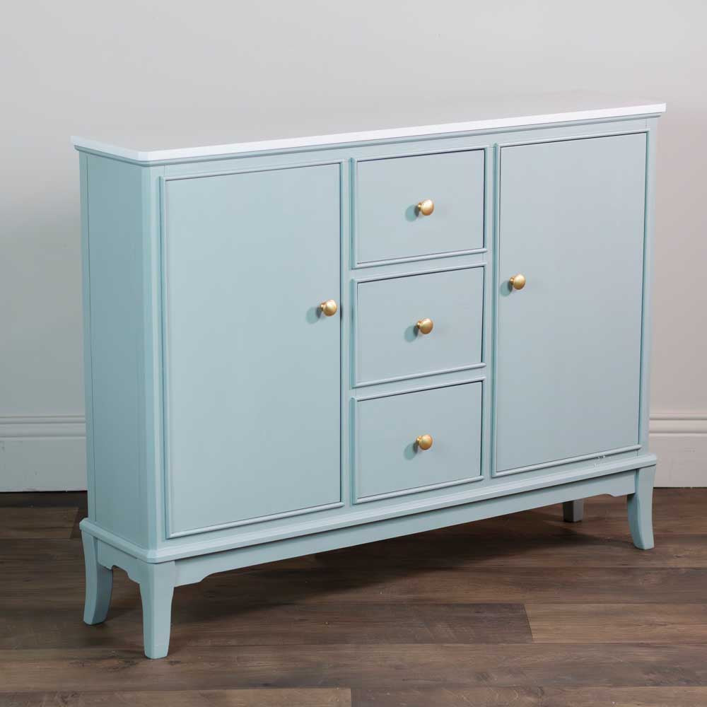 A narrow sage green sideboard with three drawers and two doors, accented with small gold knobs, set against a neutral wall and dark wood floor.