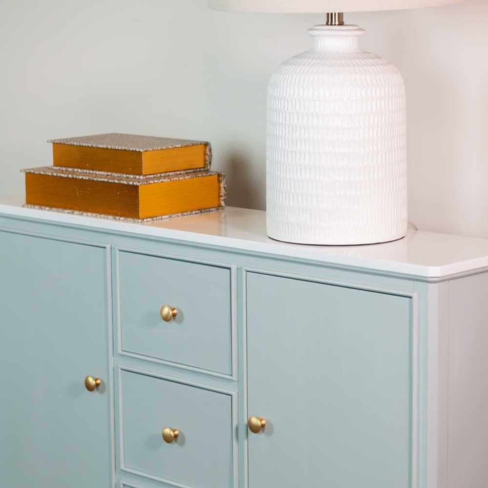 A sage green sideboard with gold knobs and a white marble top surface, styled with two stacked gold books and a large white textured table lamp against a pale wall.