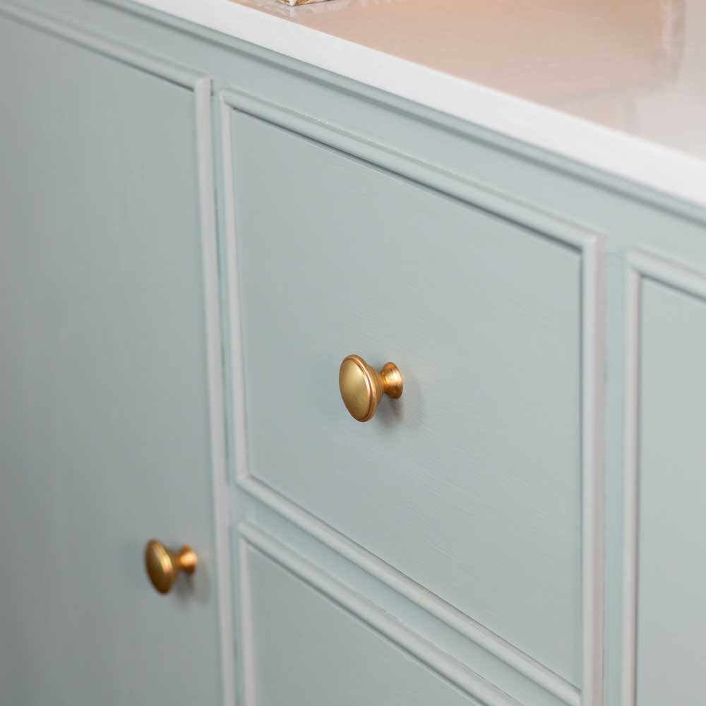 A close-up of a light sage green sideboard with a white marble countertop, featuring two round brass drawer and cabinet knobs.