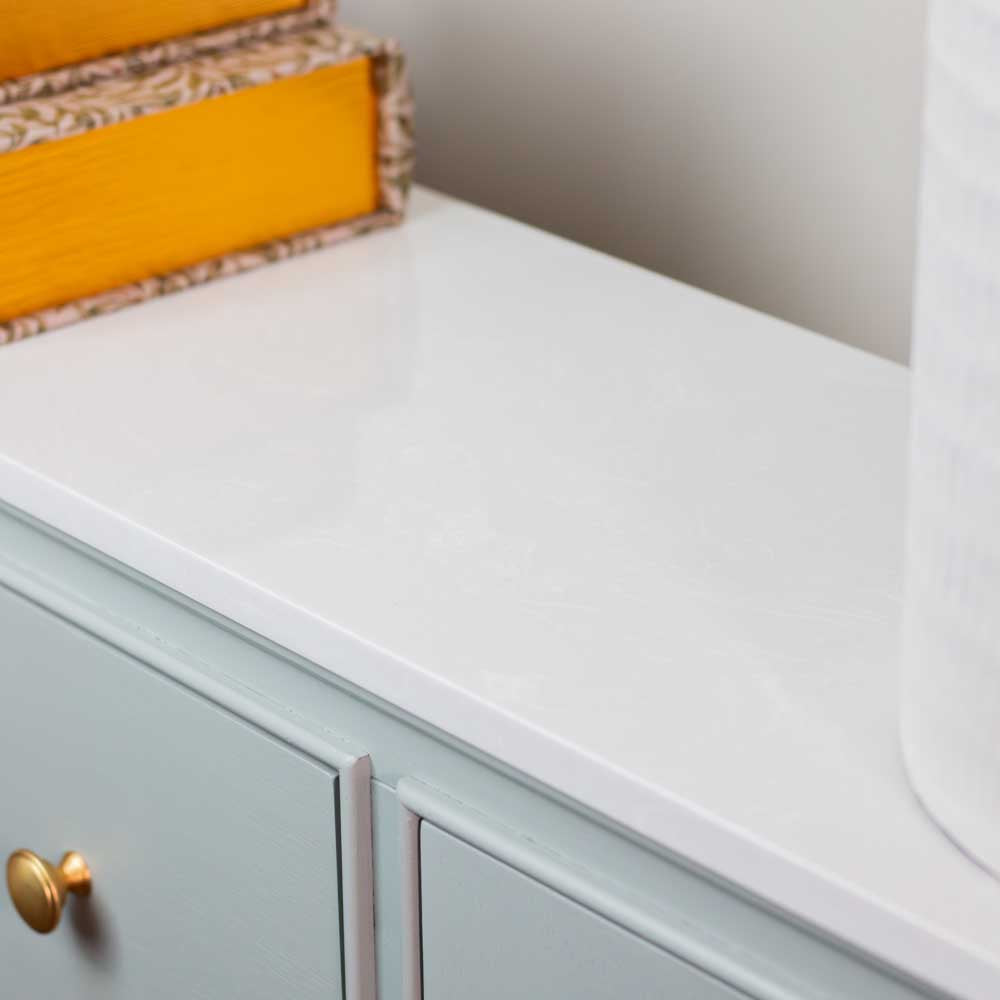 A close-up of a glossy white marble countertop with a sage green sideboard below, featuring a gold round knob, with decorative books and a white vase partially visible.