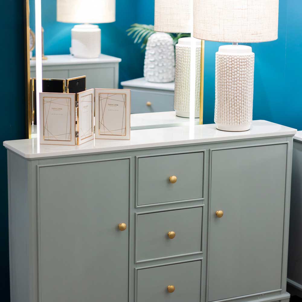 A sage green wooden sideboard or dresser with three drawers and two cabinet doors, accented with round gold knobs, styled with white textured table lamps and picture frames against a bright blue wall.