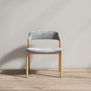 A modern dining chair with a light gray velvet upholstered seat and curved backrest, supported by a natural light-wood frame, set against a neutral gray wall and light wood floor.