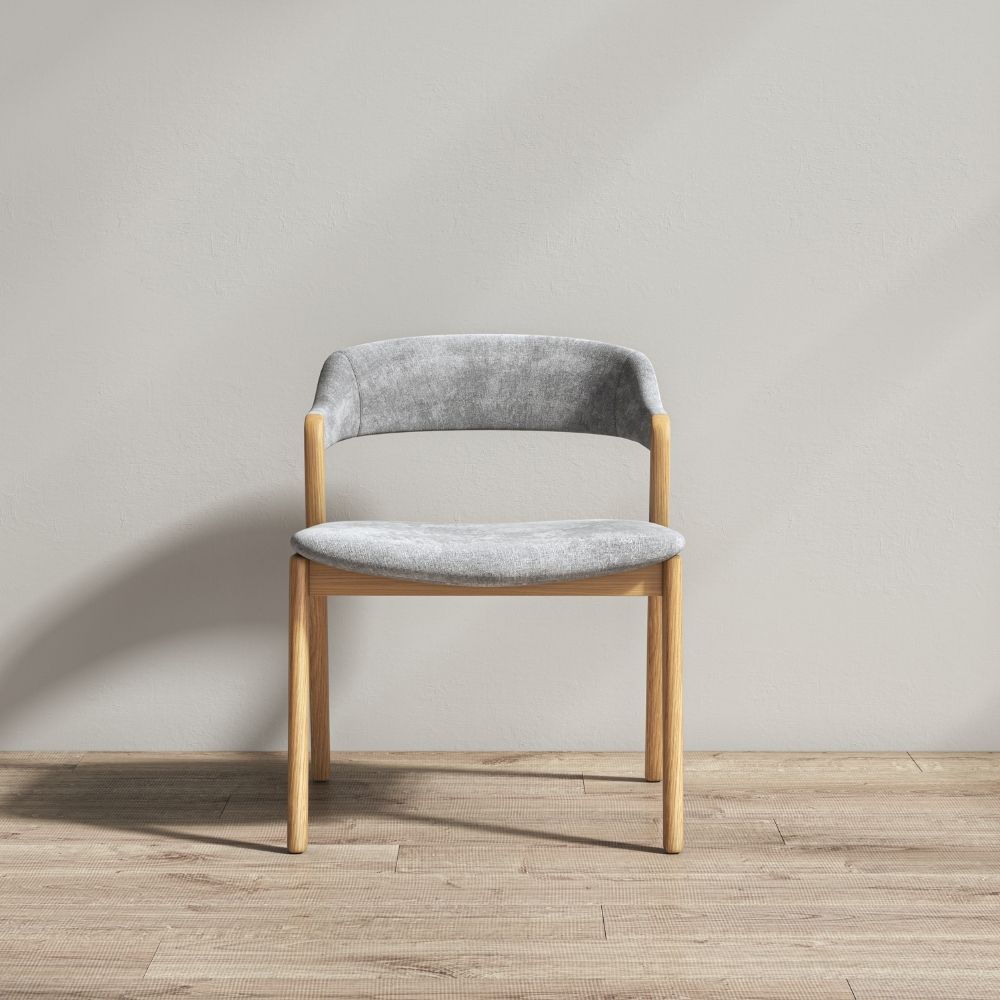 A modern dining chair with a light gray velvet upholstered seat and curved backrest, supported by a natural light-wood frame, set against a neutral gray wall and light wood floor.